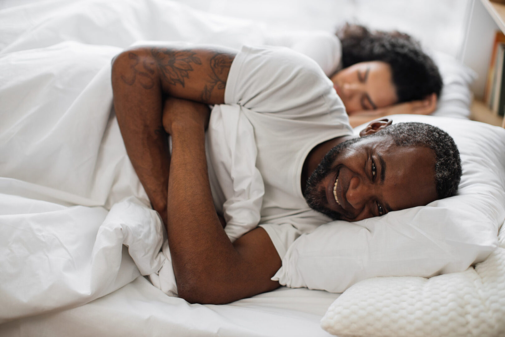 Smiling bearded husband relaxing on bed with pillow under head while sleeping wife resting on background. Cheerful african american man in mature relationship relishing full life.